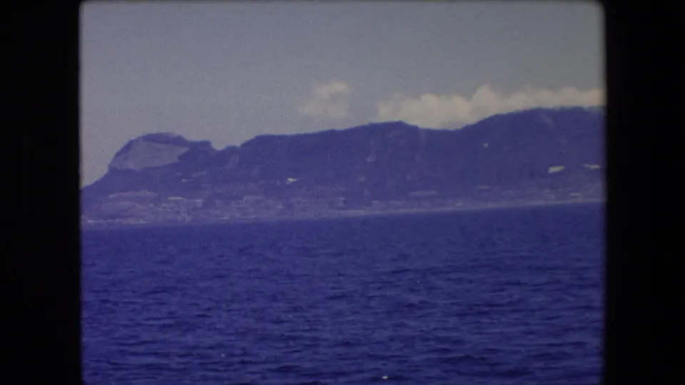 1971 Barcelona-area 8mm travel frame showing a distant mountainous coast or island beyond dark blue water, with pale clouds above and dark film borders around the frame.