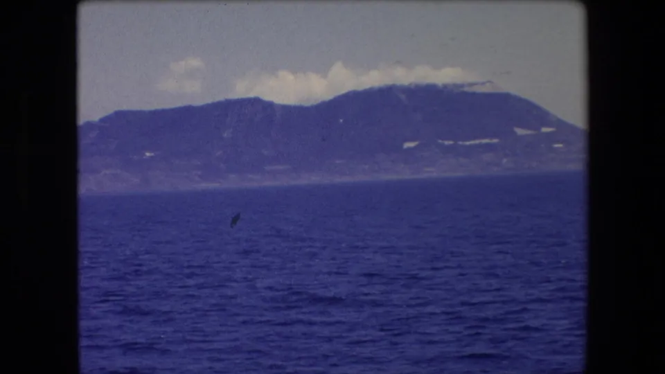 1971 Barcelona-area 8mm frame with a broad rounded mountain or coastal ridge across the sea, scattered cloud cover above, and a small bird-like speck visible near the middle distance.