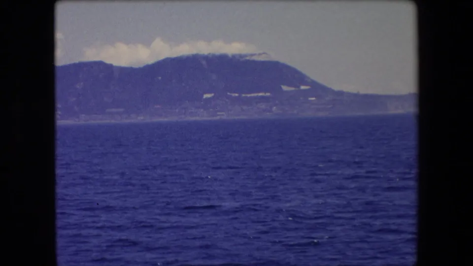 1971 Barcelona-area 8mm travel frame looking across rippled blue water toward a distant mountain slope, hazy shoreline, and pale cloud bank near the ridge.