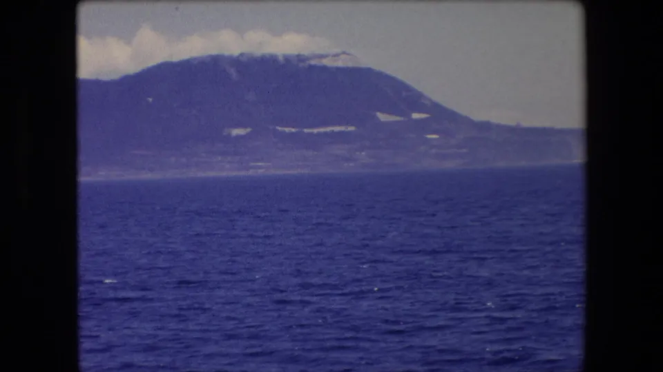 1971 Barcelona-area 8mm frame showing a large rounded mountain or peninsula across the blue sea, with light-toned patches on the slope and soft clouds along the skyline.