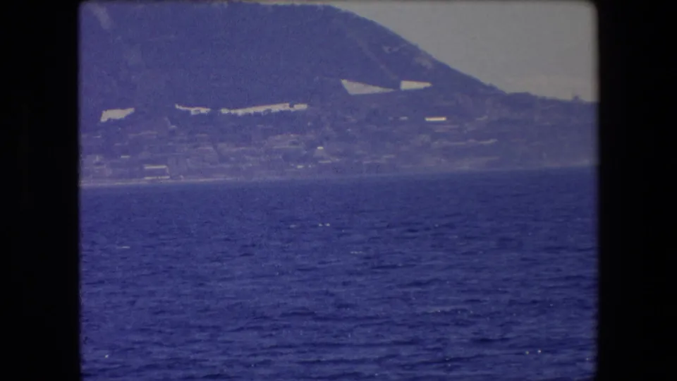 1971 Barcelona-area home-movie view of a mountainous shoreline seen across blue water from a moving boat, with pale buildings scattered along the slopes.
