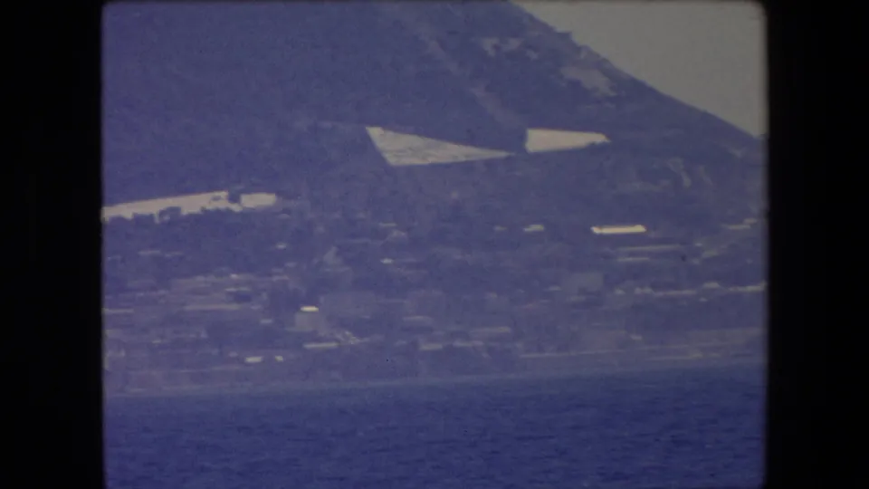 Closer home-movie view of a coastal hillside above the water, with pale structures and fields or terraces visible through a blue film cast.