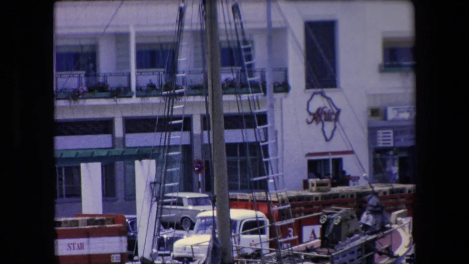 1971 Barcelona waterfront frame showing dense boat rigging and ladders before the Residencia Africa building, with a parked car and dockside equipment at street level.