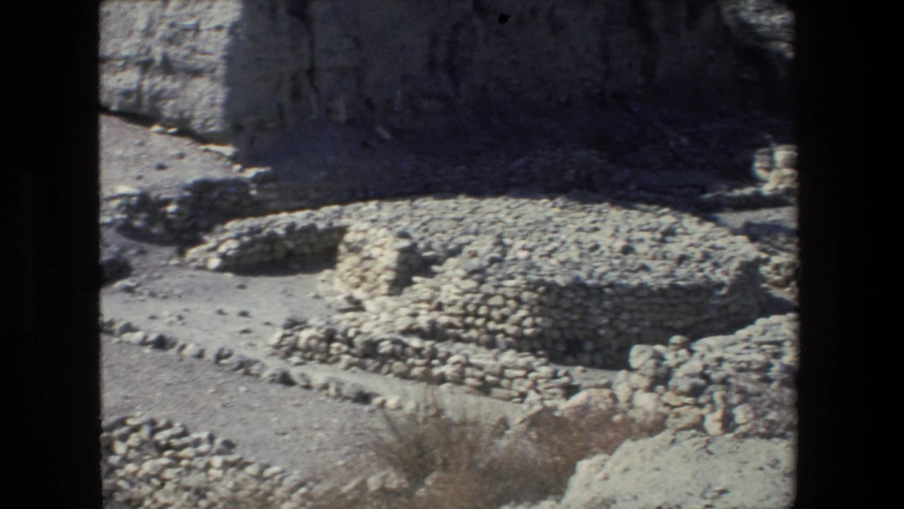 Circular low stone ruin beside a rock wall in bright sun.