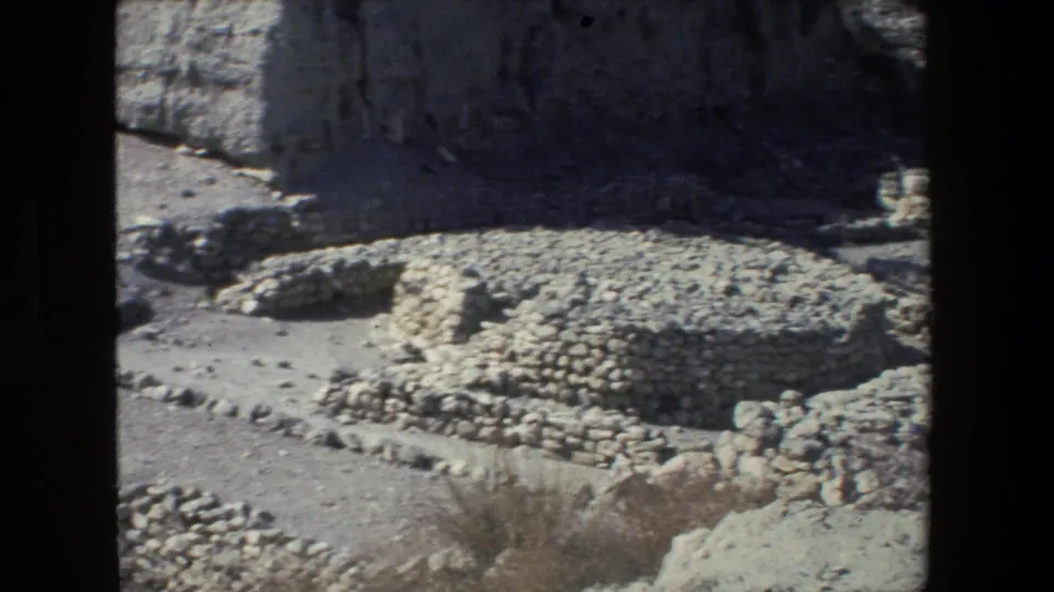 1972 Israel 8mm frame showing low circular stone structures on dry ground beneath a pale rock wall, filmed from a visitor viewpoint.