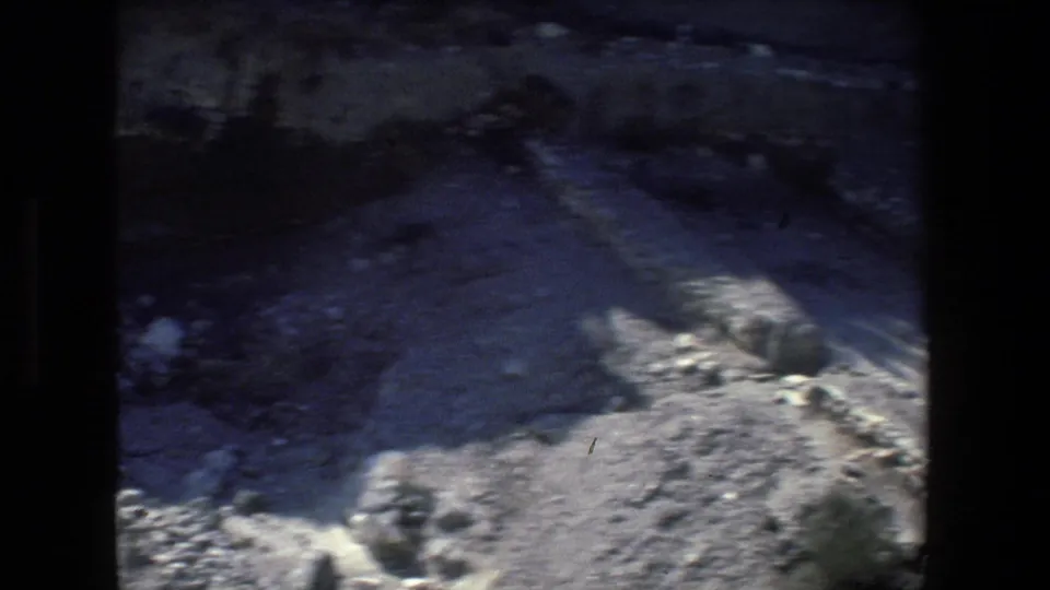 1972 Israel 8mm frame showing a shadowed rocky depression with pale stone edges, rough canyon surfaces, and scattered rocks under strong sunlight.