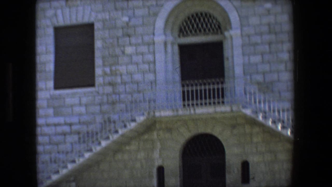A pale stone facade with an arched doorway, barred upper opening, stairway, and metal railings fills the frame in this 1972 8mm Tel Aviv, Israel clip.