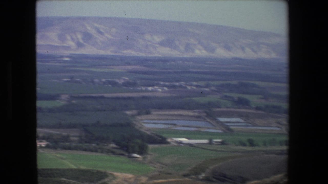 Aerial 8mm frame near Tel Aviv with green fields, rectangular ponds or reservoirs, scattered low structures, and arid hills beyond the plain.