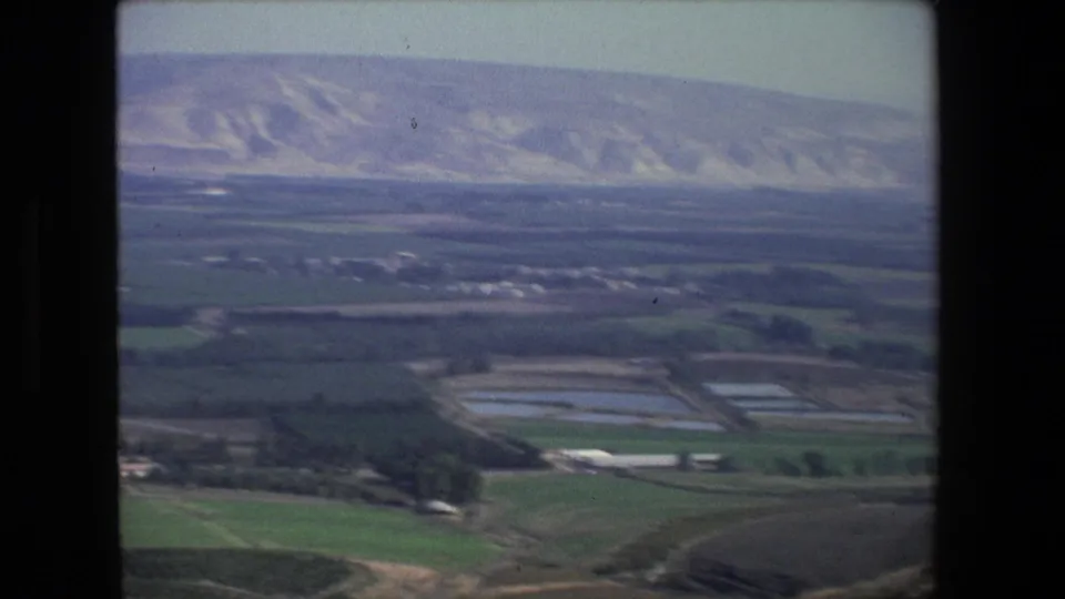 Aerial 8mm frame near Tel Aviv with green fields, rectangular ponds or reservoirs, scattered low structures, and arid hills beyond the plain.