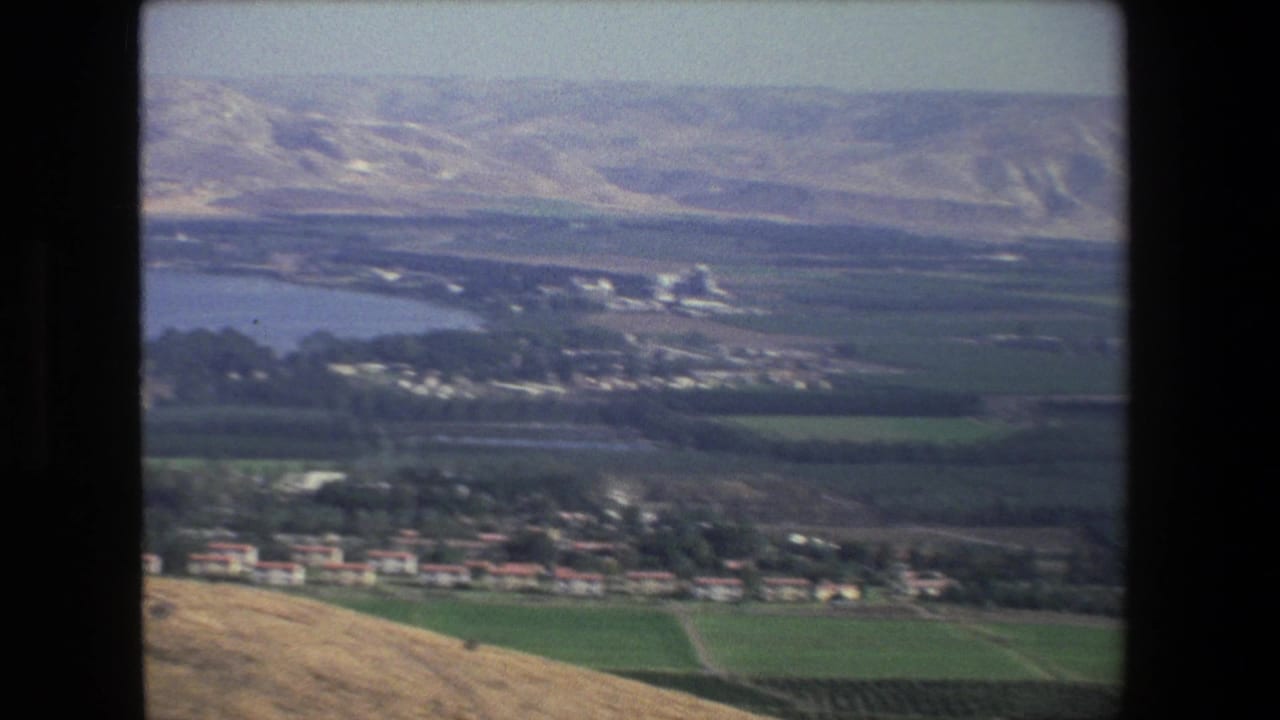 1972 aerial-style home-movie frame near Tel Aviv showing a low settlement edge, green fields, a body of water, and pale hills in the background.