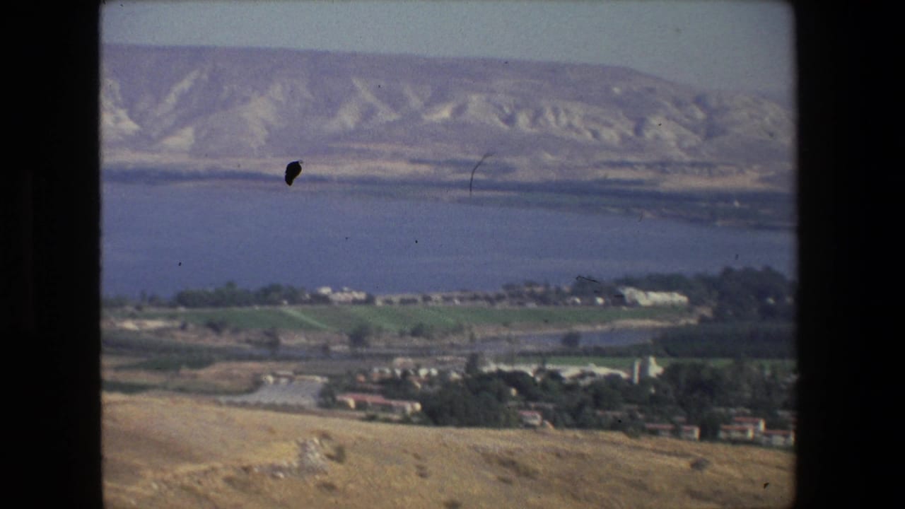 Aerial 8mm frame near Tel Aviv showing shoreline water, agricultural fields, low settlement buildings, roads, and pale hills across the distance.