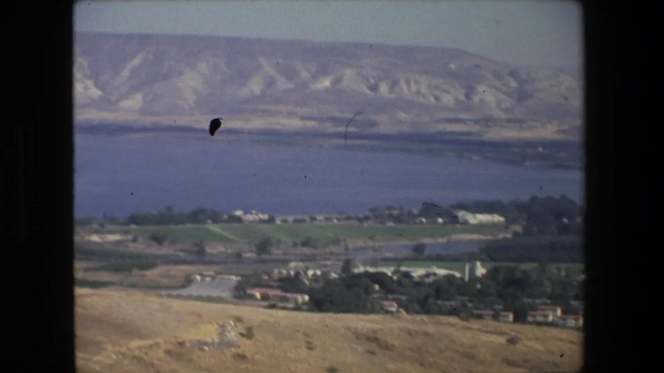 Aerial 8mm frame near Tel Aviv showing shoreline water, agricultural fields, low settlement buildings, roads, and pale hills across the distance.