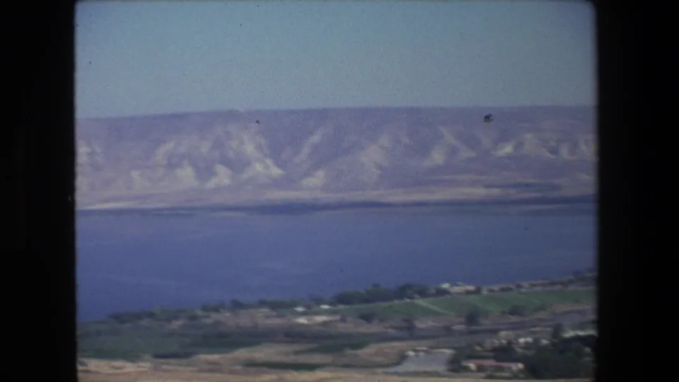 1972 8mm travel frame of an Israeli waterside panorama, with blue water, distant pale hills, foreground greenery, and dark projector-like borders.