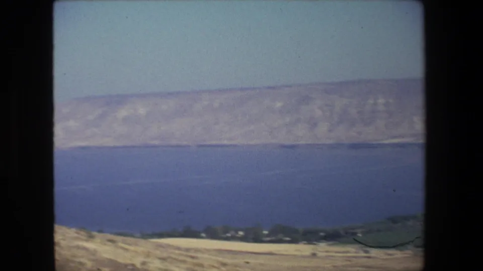 1972 8mm travel frame looking across a large blue body of water toward dry hills and a low shoreline in Israel, with soft focus and film grain.