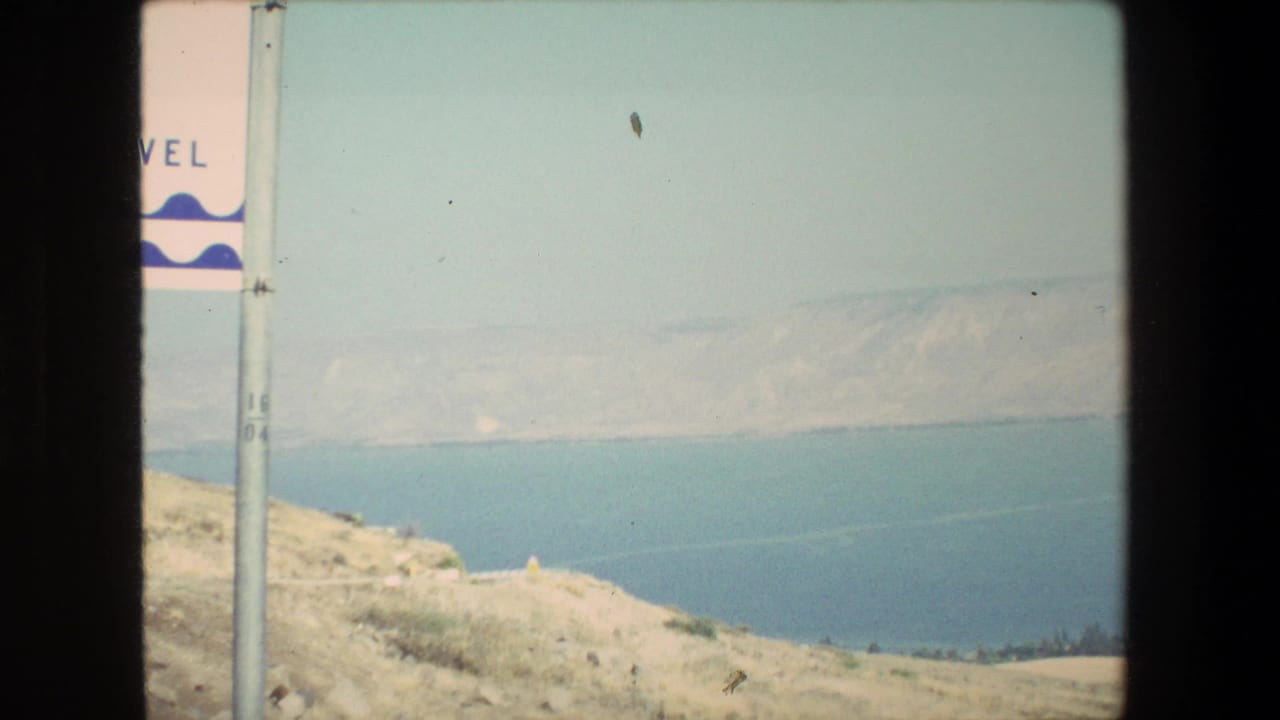 1972 8mm travel frame with a partial roadside sign at left, pale hillside foreground, blue water beyond, and distant hills under hazy light.