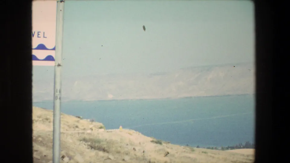 1972 8mm travel frame with a partial roadside sign at left, pale hillside foreground, blue water beyond, and distant hills under hazy light.