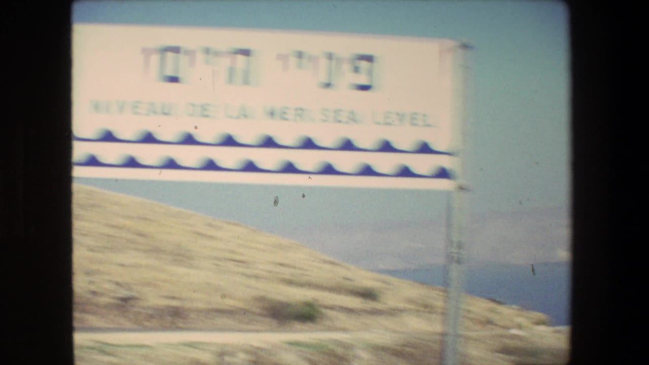 1972 8mm travel frame showing a roadside sign with Hebrew and English text, including visible wording that appears to refer to the Kinneret, set against dry hills and blue water.