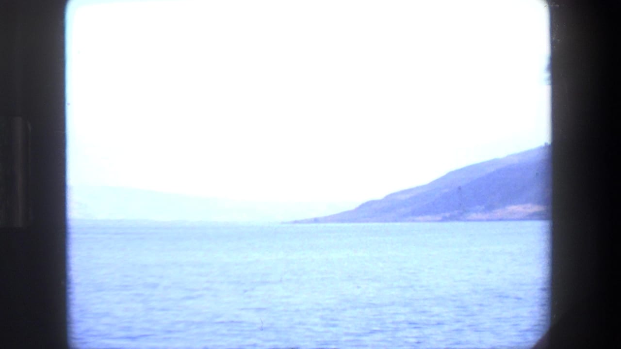 1972 Tel Aviv travel-film frame showing calm blue water, a hazy shoreline, distant hills, and heavy dark borders from the 8mm film gate.