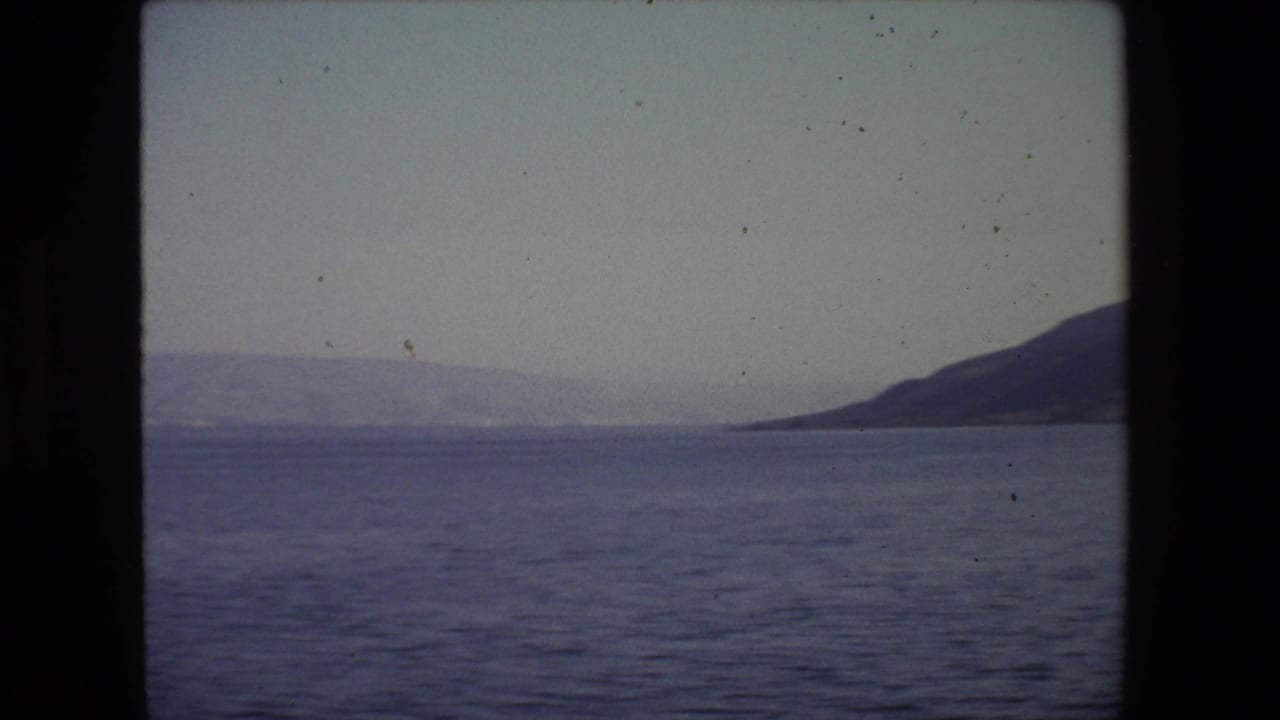 1972 Tel Aviv coastal view from a moving boat, with dark 8mm frame edges around open water, a distant shoreline, and a low hill at right.