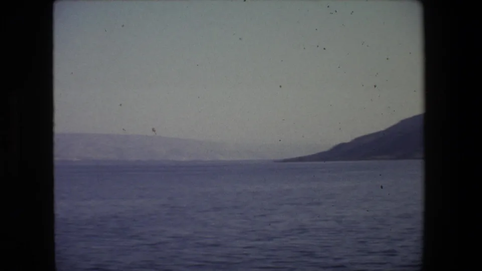 1972 Tel Aviv coastal view from a moving boat, with dark 8mm frame edges around open water, a distant shoreline, and a low hill at right.