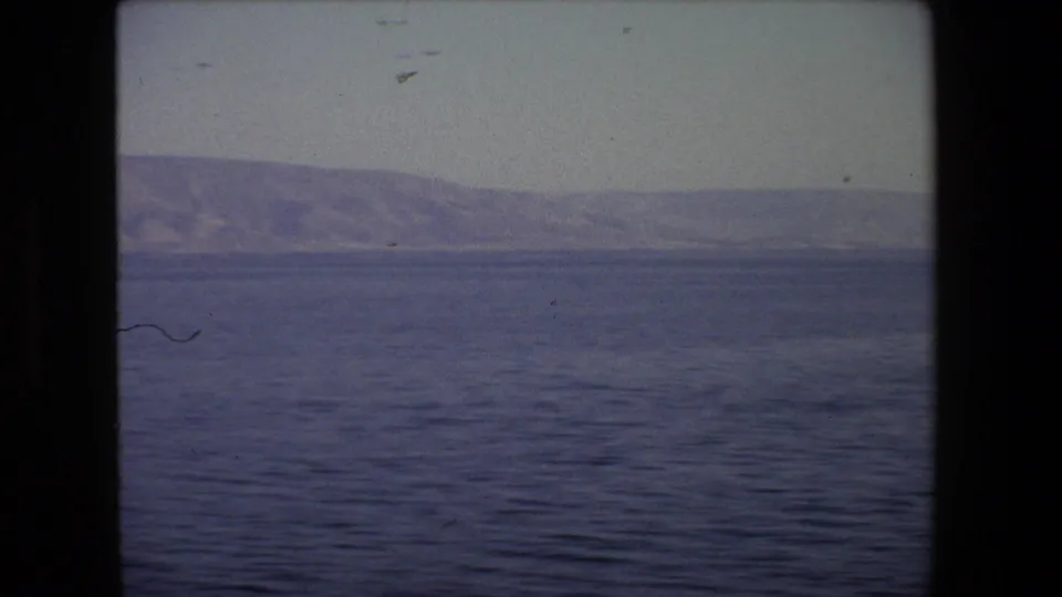 1972 Tel Aviv coastal travel frame with blue water in the foreground, a distant hazy hillside, dark 8mm borders, and small visible dust marks on the scan.