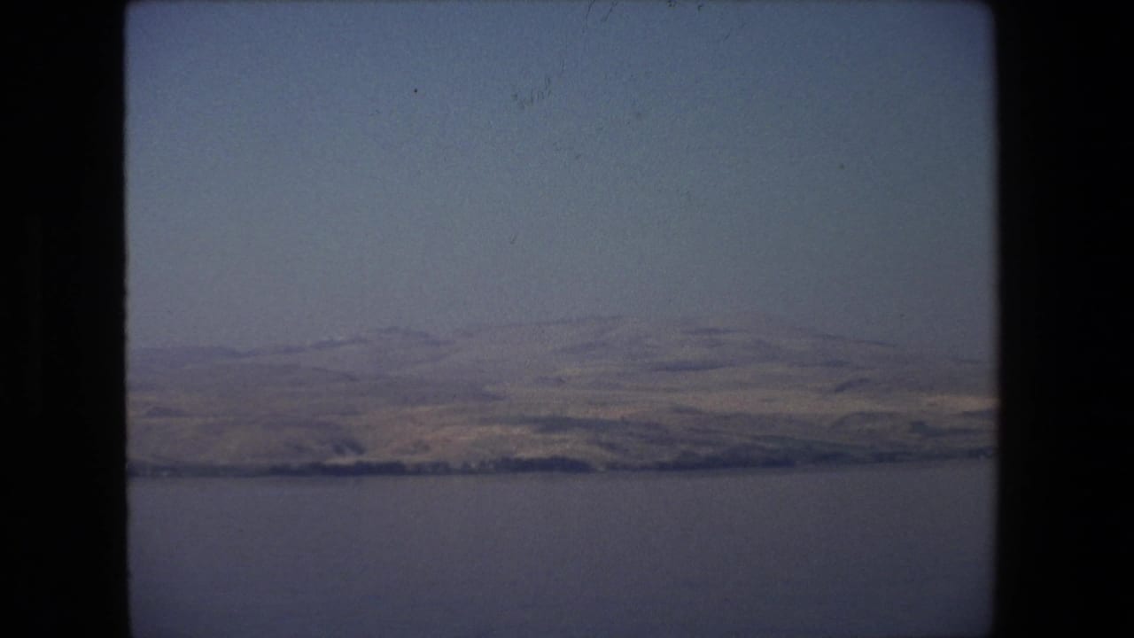 Calm water below distant dry hills under a hazy sky.