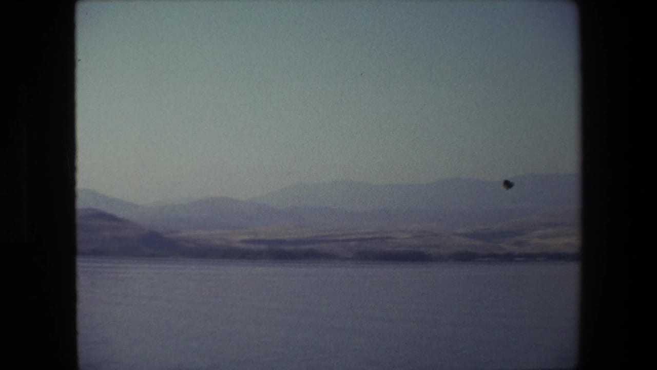 1972 Tel Aviv, Israel home-movie frame with calm water, layered distant hills, dry shoreline terrain, and a pale sky in faded 8mm color.