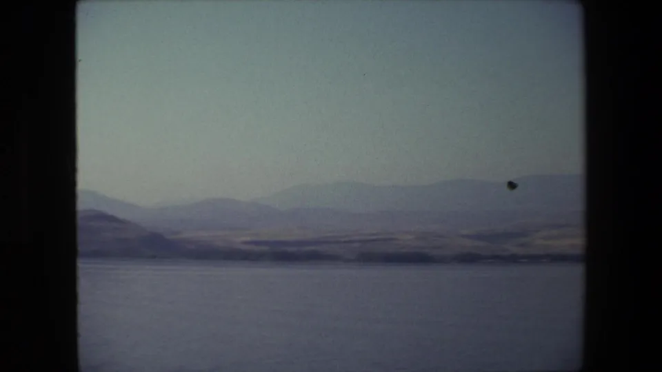 1972 Tel Aviv, Israel home-movie frame with calm water, layered distant hills, dry shoreline terrain, and a pale sky in faded 8mm color.