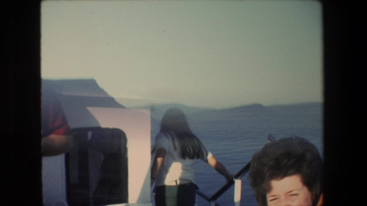 1972 Israel 8mm frame from a boat showing passengers near the rail, a seated woman partly visible in the foreground, and hazy water and hills beyond.