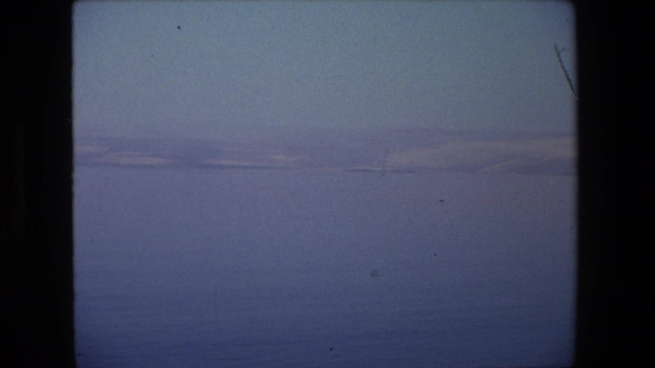 A handheld 8mm view across open blue water toward a hazy distant shoreline or mountain ridge, with no clear people centered in the frame.