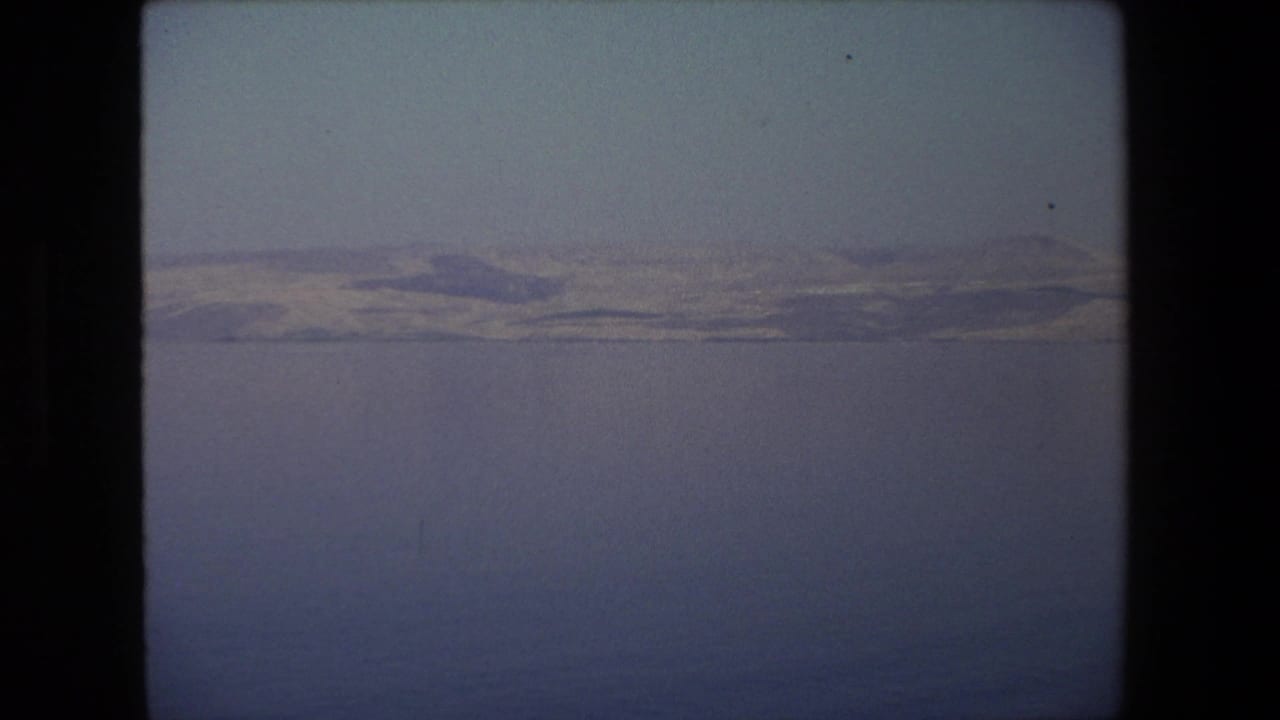 1972 home-movie landscape frame associated with Tel Aviv, Israel, with still water in the foreground and a subdued mountain ridge in the distance.
