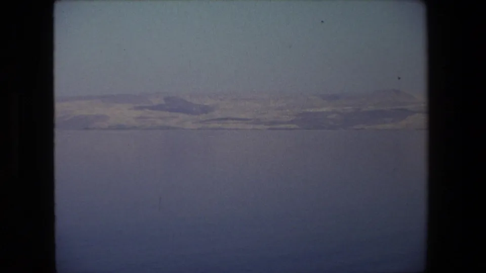 1972 home-movie landscape frame associated with Tel Aviv, Israel, with still water in the foreground and a subdued mountain ridge in the distance.