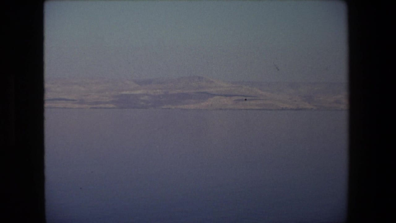 1972 8mm frame from Tel Aviv, Israel, showing calm water in the foreground and low arid hills stretching along a distant shoreline.