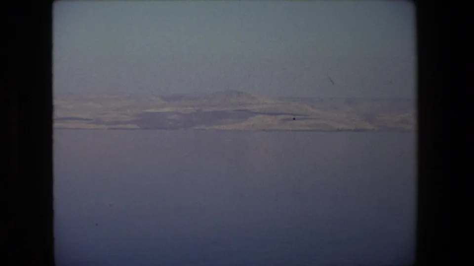 1972 8mm frame from Tel Aviv, Israel, showing calm water in the foreground and low arid hills stretching along a distant shoreline.