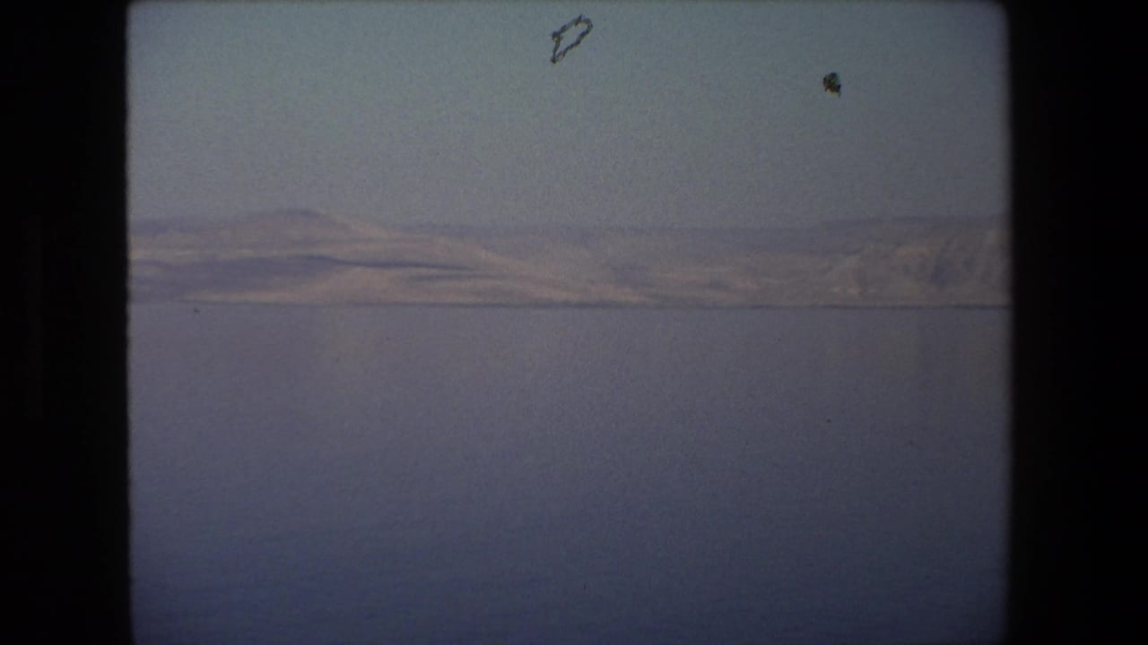 1972 8mm frame from Tel Aviv, Israel, with still blue water below a thin shoreline and muted hills in hazy light.