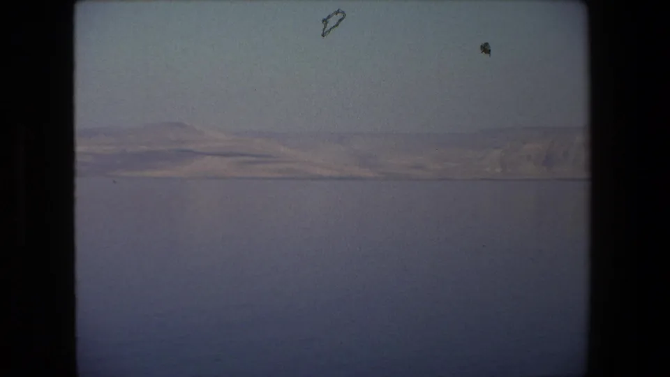 1972 8mm frame from Tel Aviv, Israel, with still blue water below a thin shoreline and muted hills in hazy light.