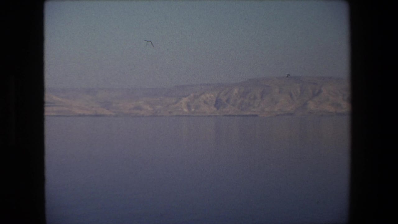 1972 8mm frame from Tel Aviv, Israel, showing rugged sunlit hills meeting a narrow shoreline beyond calm water.