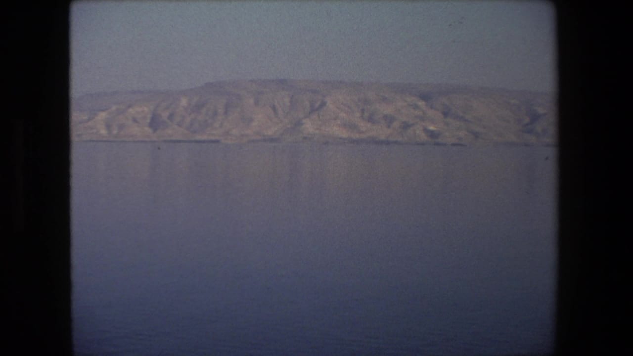 1972 Tel Aviv, Israel home-movie frame showing a calm expanse of water with rugged dry hills on the far horizon and soft 8mm vignetting.