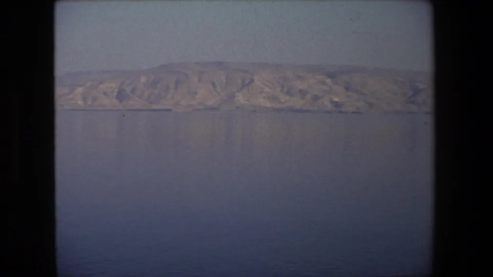 1972 Tel Aviv, Israel home-movie frame showing a calm expanse of water with rugged dry hills on the far horizon and soft 8mm vignetting.