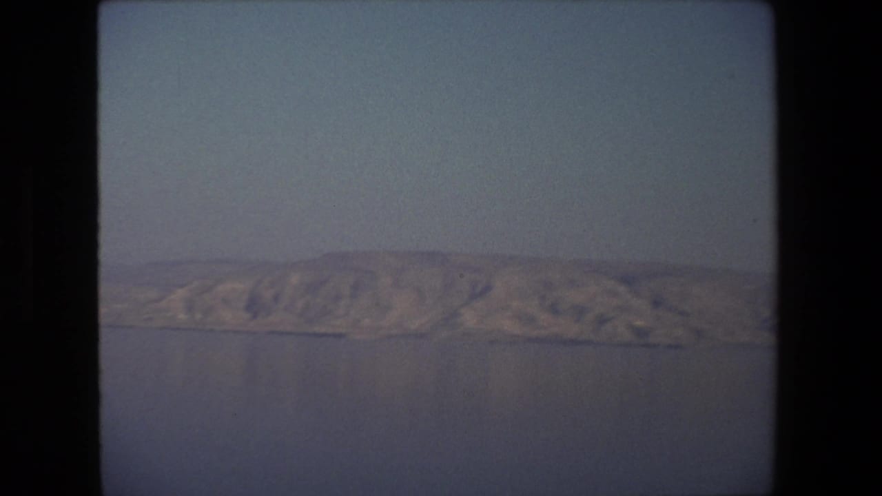 1972 Tel Aviv, Israel 8mm frame showing rugged sunlit hills across calm water, with haze, grain, and dark contact-sheet borders visible.
