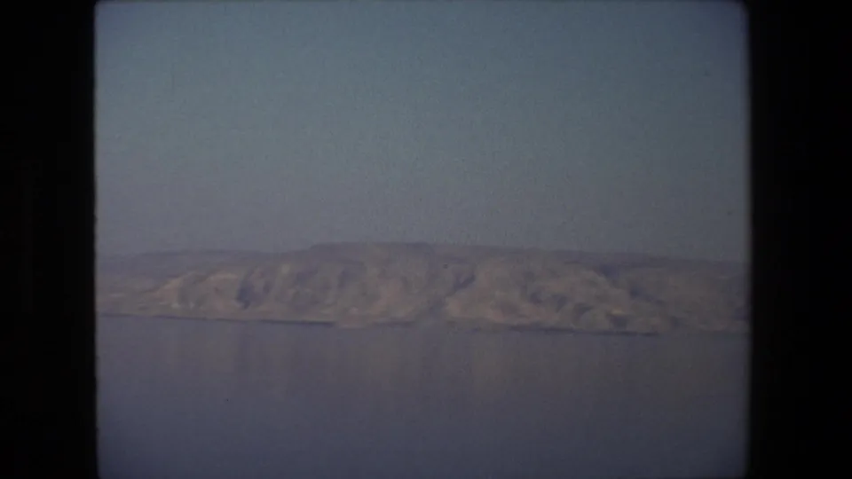 1972 Tel Aviv, Israel 8mm frame showing rugged sunlit hills across calm water, with haze, grain, and dark contact-sheet borders visible.
