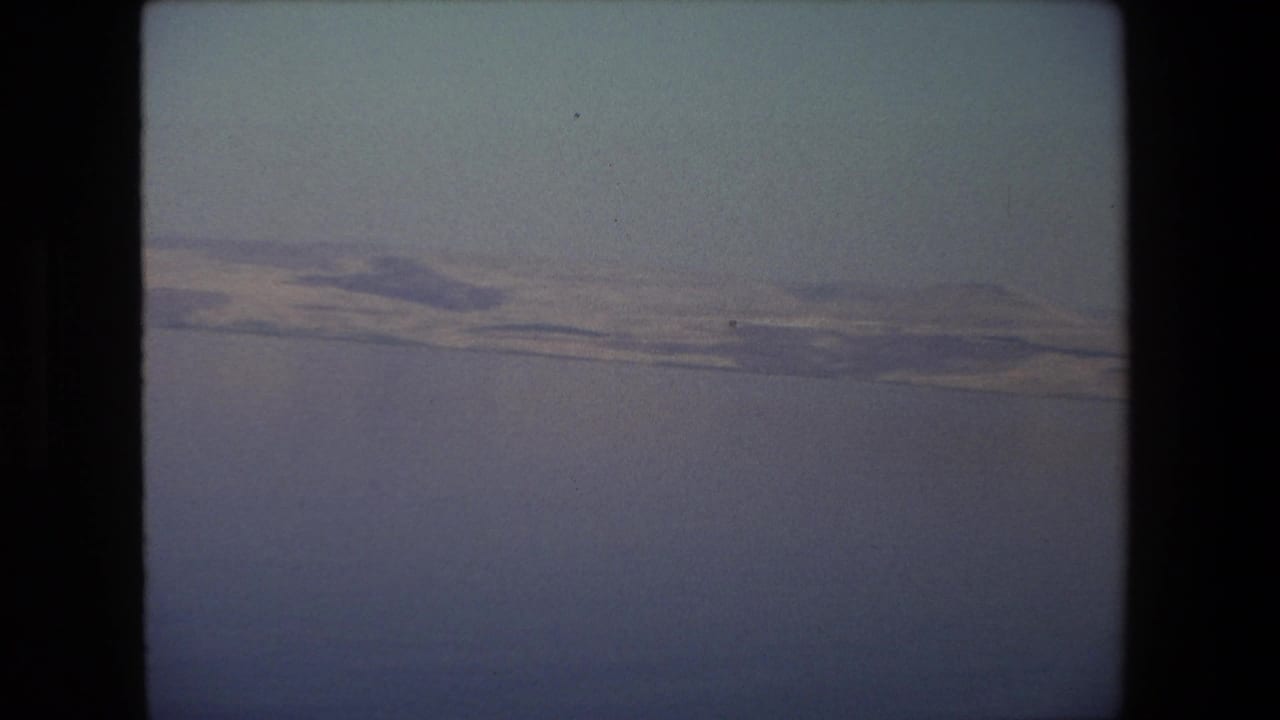 1972 Tel Aviv-area 8mm frame with a broad expanse of water, a pale distant embankment, and low hills softened by haze.