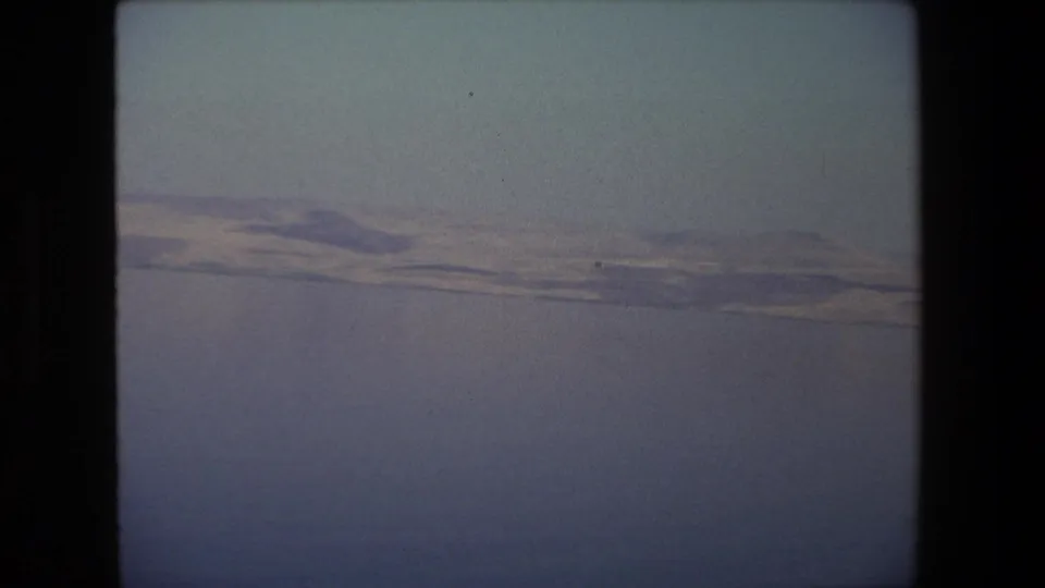 1972 Tel Aviv-area 8mm frame with a broad expanse of water, a pale distant embankment, and low hills softened by haze.