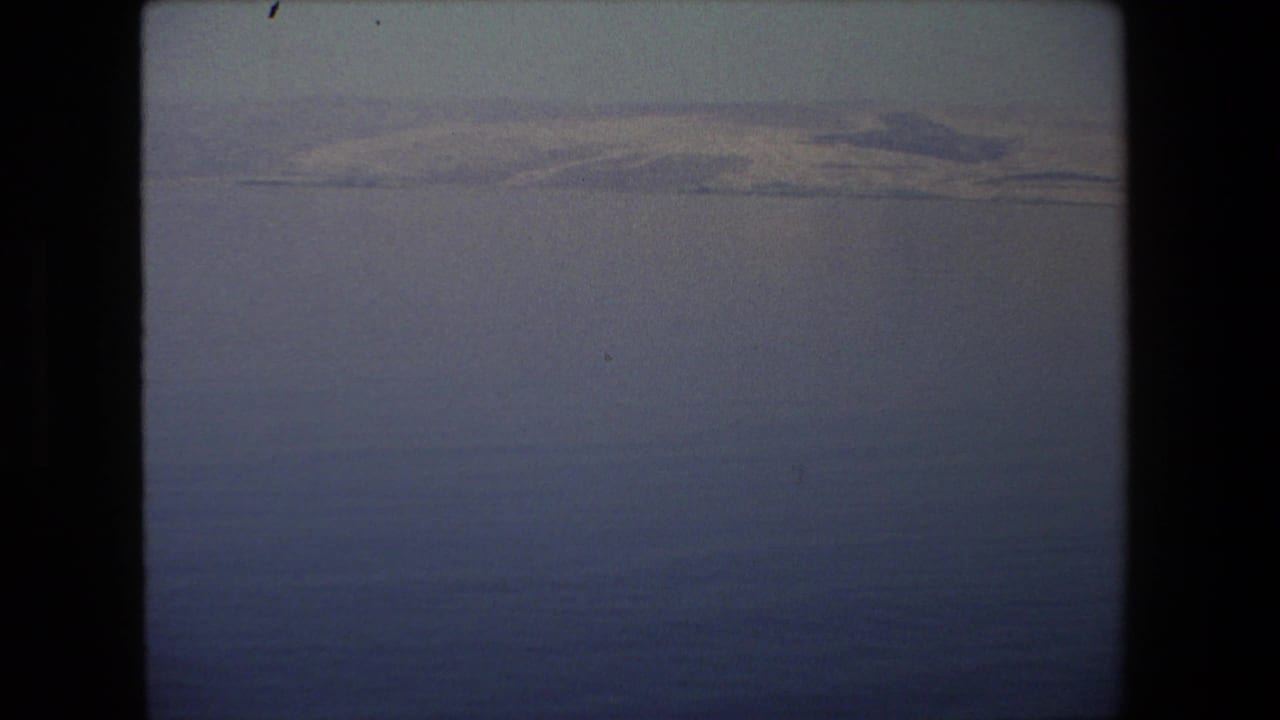 1972 Tel Aviv-area 8mm frame looking over dark blue water toward a low, pale shoreline with faint hills and visible home-movie grain.