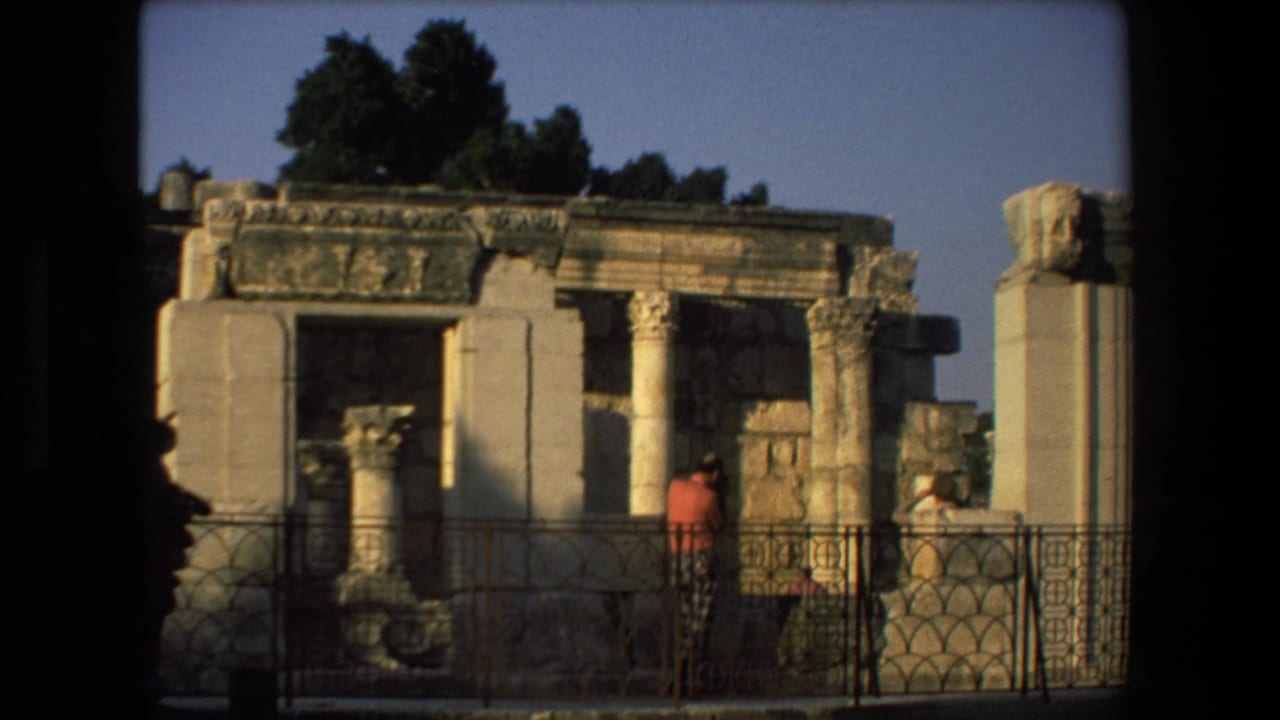 Tourists at an ancient stone ruin site in Israel, with weathered columns, carved masonry, trees, and a metal viewing railing visible.