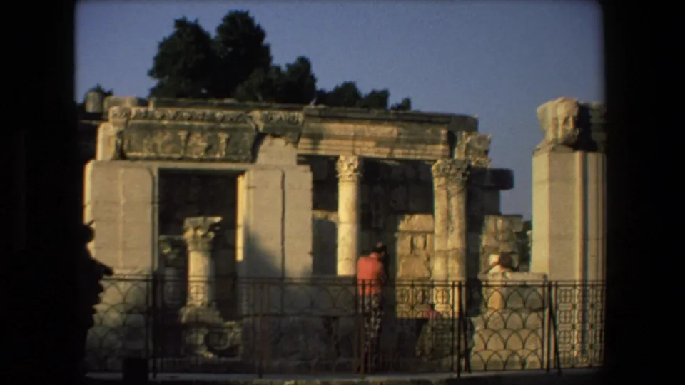Tourists at an ancient stone ruin site in Israel, with weathered columns, carved masonry, trees, and a metal viewing railing visible.
