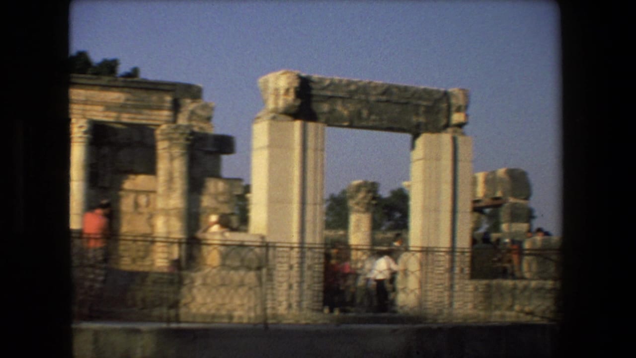 A handheld 8mm view across ancient ruins in Israel, showing broken columns, a stone lintel, metal railings, and several tourists in casual clothing.