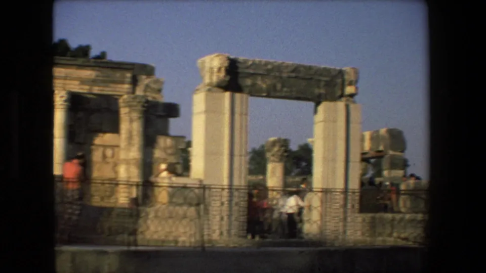 A handheld 8mm view across ancient ruins in Israel, showing broken columns, a stone lintel, metal railings, and several tourists in casual clothing.