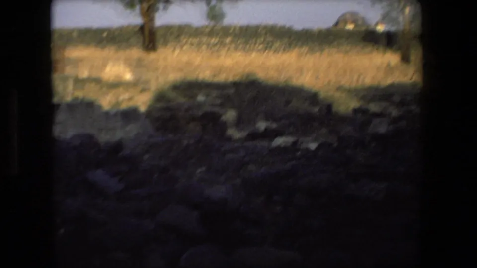 Handheld 8mm view across rocky ground, dry grass, and distant trees at the Tel Aviv ruin site, with heavy dark frame edges.