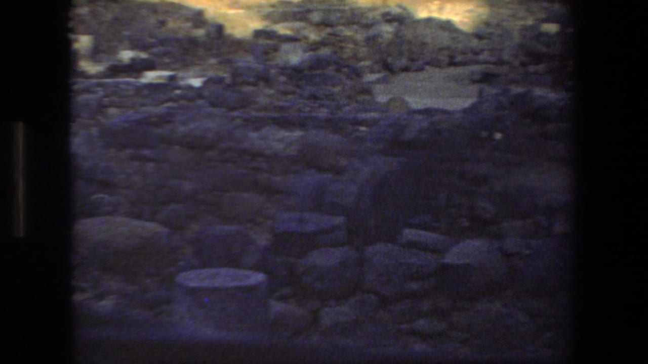 1972 home-movie frame looking down over rough stone rubble and ruin fragments, with blue-shadowed film color and dark scan borders.